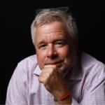 Portrait of a middle-aged man with gray hair in a pink checkered shirt, resting his chin on his hand.