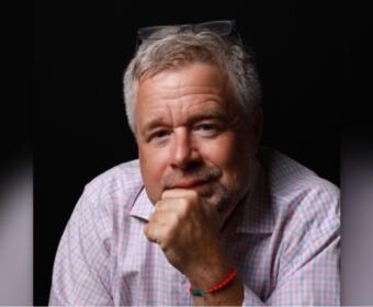 Portrait of a middle-aged man with gray hair in a pink checkered shirt, resting his chin on his hand.