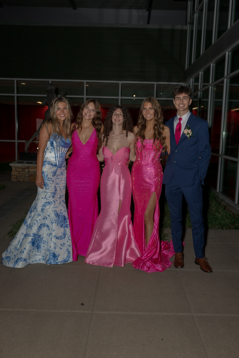 Five high school students in formal wear pose together outside a modern building at night, smiling for a group photo.