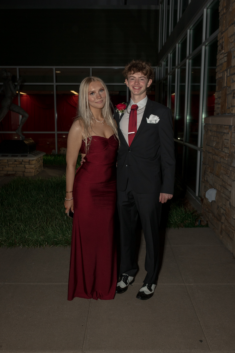 A young couple in formal attire posing outside a modern building: woman in a burgundy gown and man in a black suit with a red tie and white pocket square.