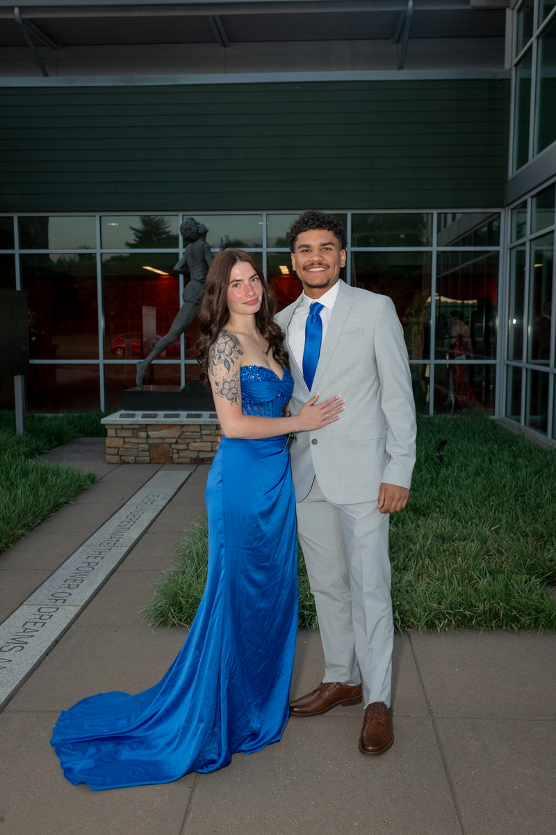Couple in formal attire posing outside a building; woman in a blue evening gown and train, man in a light gray suit with a blue tie, smiling.