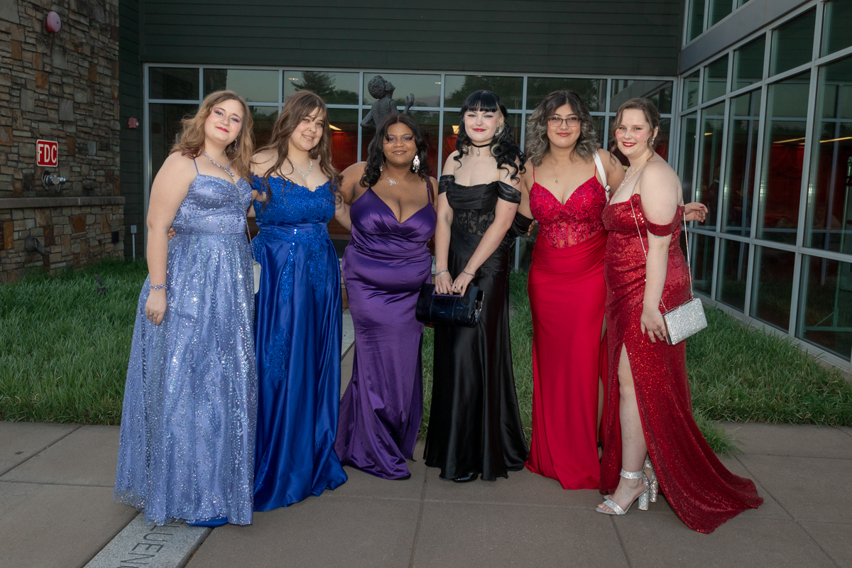 Six young women in formal evening gowns pose outside a building for a group photo at a formal event.