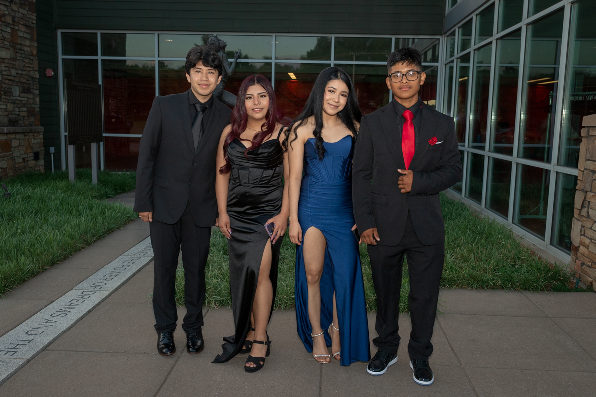 Four young adults in formal attire pose outside a modern glass building, two men in black suits and two women in high-slit gowns (black and blue).