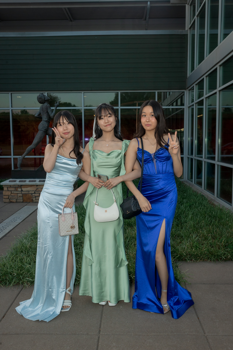 Three women in satin evening gowns pose outdoors, each flashing a peace sign. Online backdrop: glass-front building behind them.