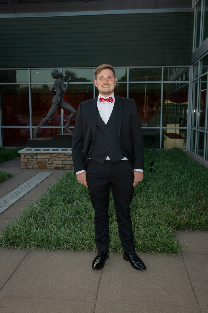 Young man in a black tuxedo with a red bow tie and vest stands on a sidewalk in front of a glass-front building and statue sculpture.