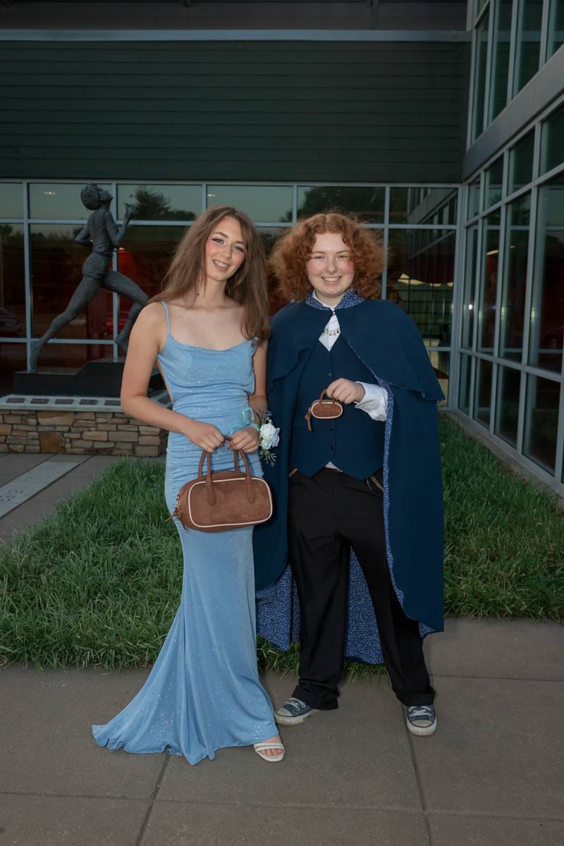 Two teens in formal wear pose outside a glass-front building; girl in a light blue gown with a brown handbag, boy in a blue cape and vest beside her.