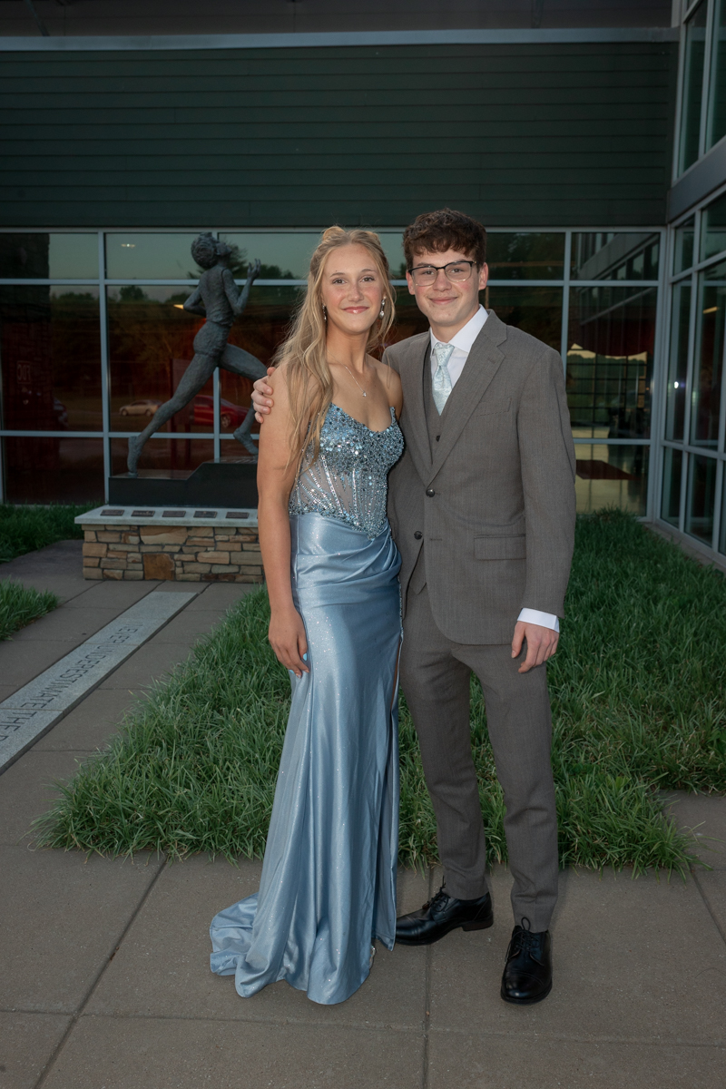 Young couple in formal attire posing outside a modern building; girl in a sparkly blue gown, boy in a gray suit.