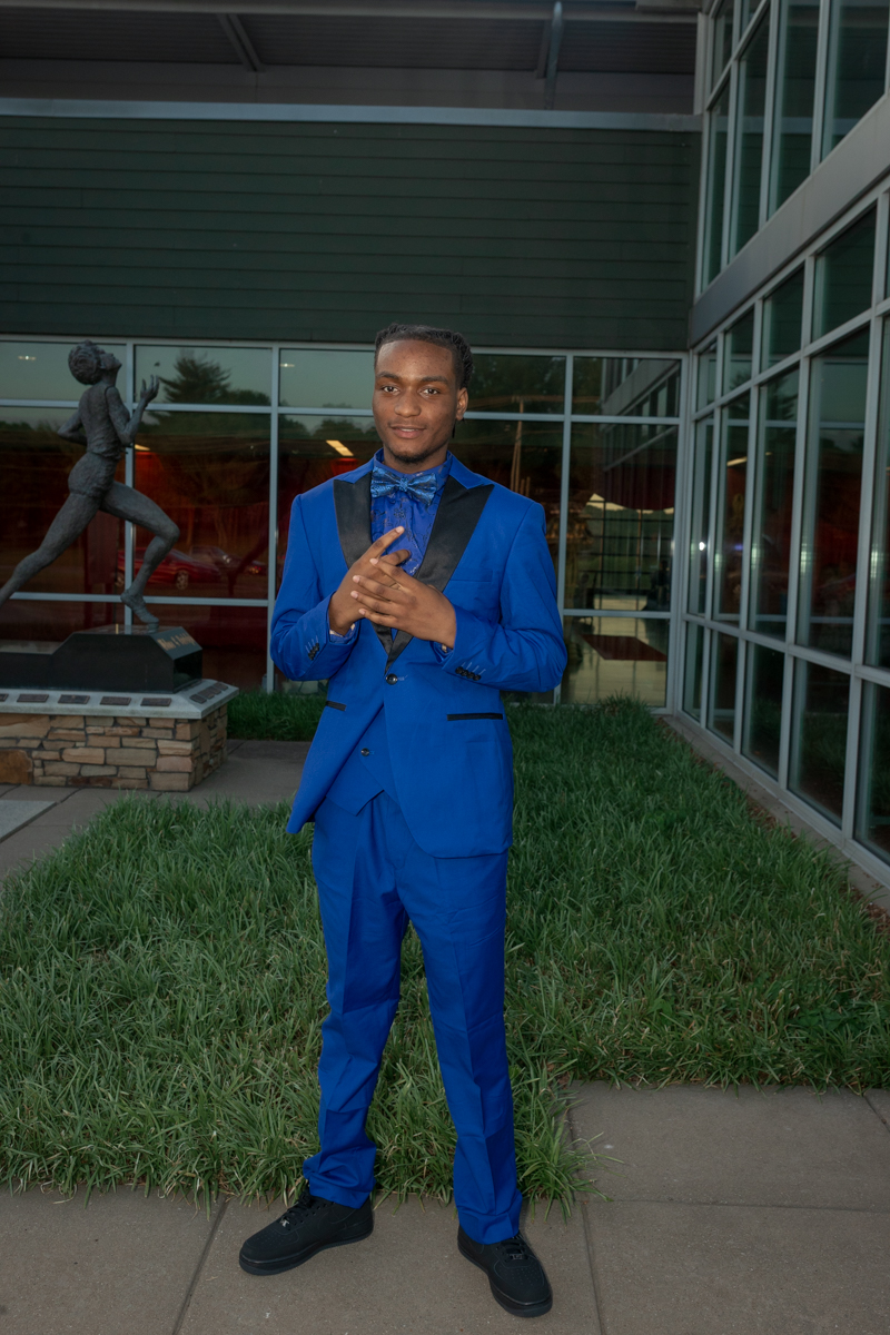 Young man in a bright blue tuxedo with a bow tie stands on a sidewalk outside a glass-front building with a statue nearby.