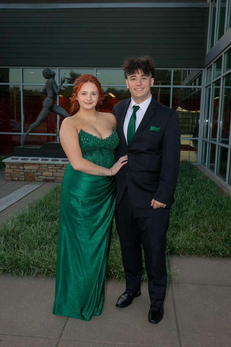 Couple in formal wear posing arm-in-arm outside a modern glass-fronted building, smiling at the camera.