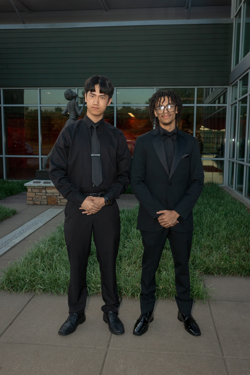 Two men in black formalwear standing side by side outdoors in front of a glass-front building.