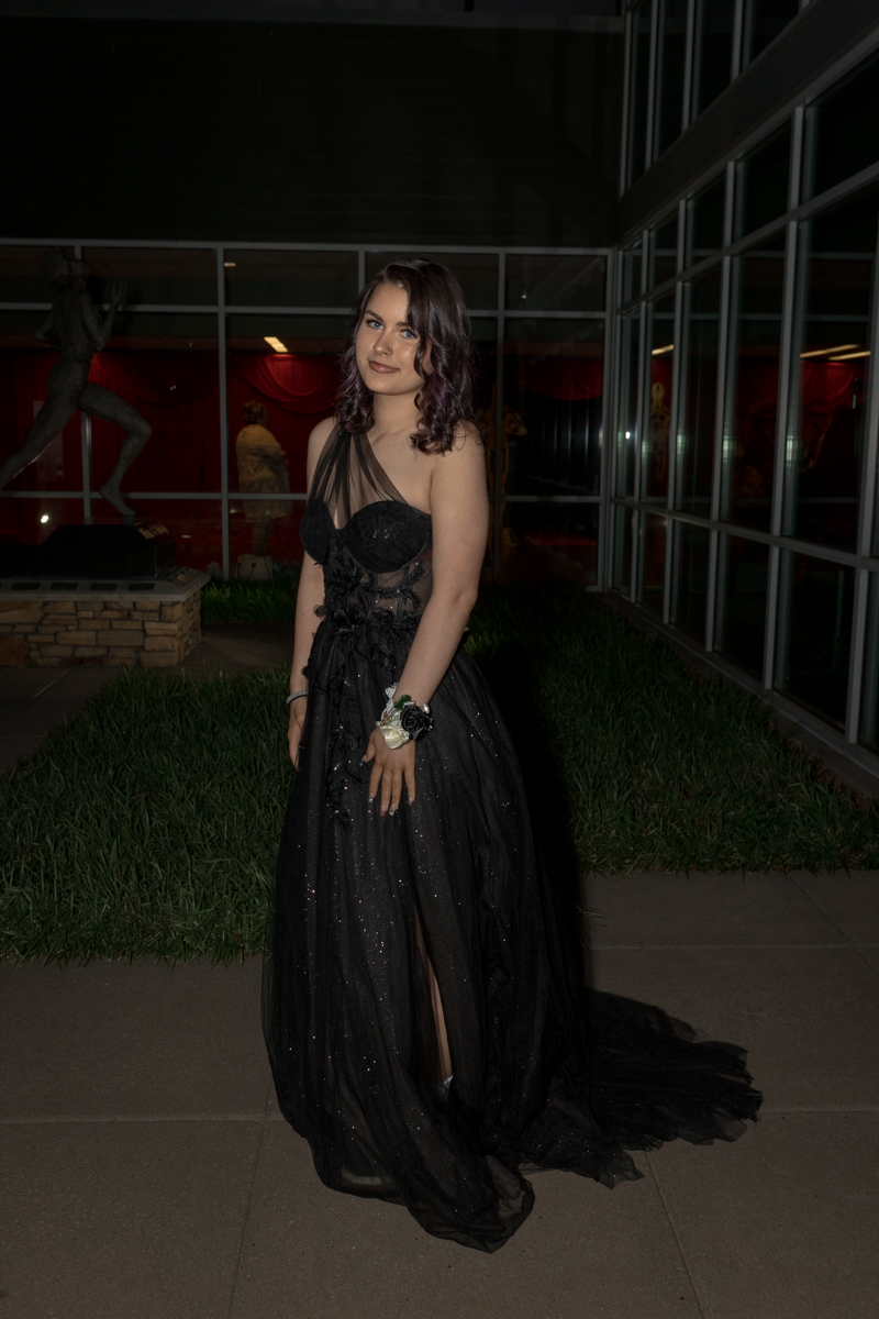 Young woman in a shimmering black evening gown with an asymmetrical sheer neckline, posing outside at night.