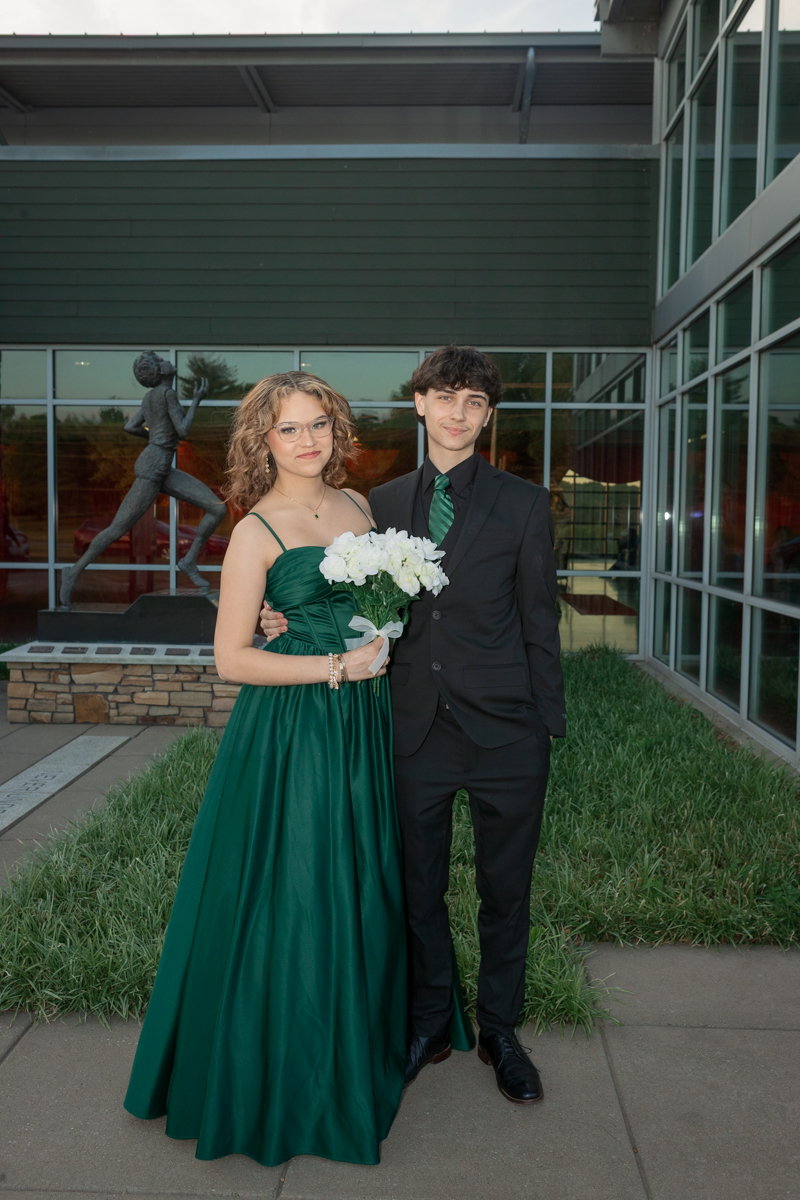 Young couple dressed for a formal event pose outside a modern building; woman in an emerald gown holding a white bouquet, man in a black suit with a green tie.
