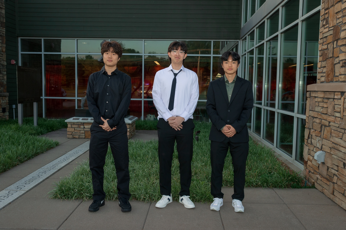 Three young men in semi-formal attire stand together outdoors in front of a modern glass-fronted building.