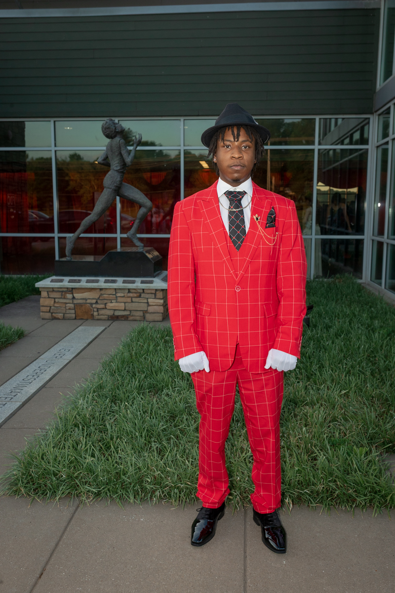Man in a bright red plaid suit with white gloves and a black fedora, standing on a sidewalk outside a glass-front building beside a statue.