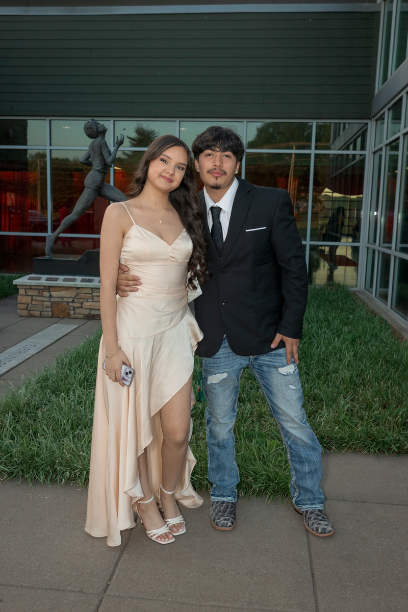 Couple posing together outside a modern glass building: woman in a peach flowing dress and heels, man in a black suit jacket and jeans with a tie.