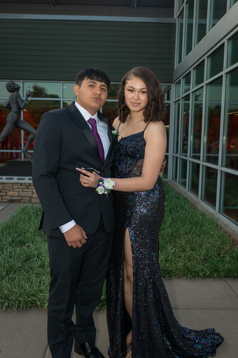 Two young people in formal attire pose arm-in-arm outside a glass-front building, the woman in a sequined gown with a leg slit and corsage, the man in a suit and purple tie.