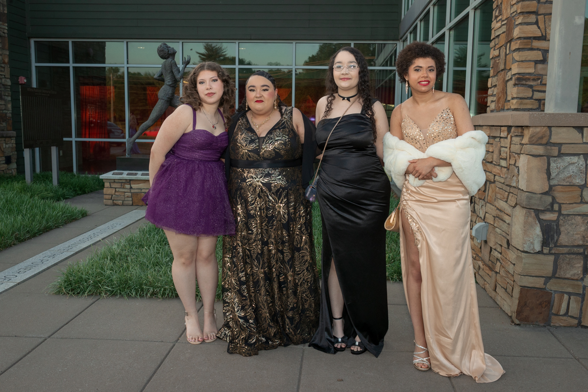 Four women in formal gowns posing together outside a modern building with large windows and stone accents.