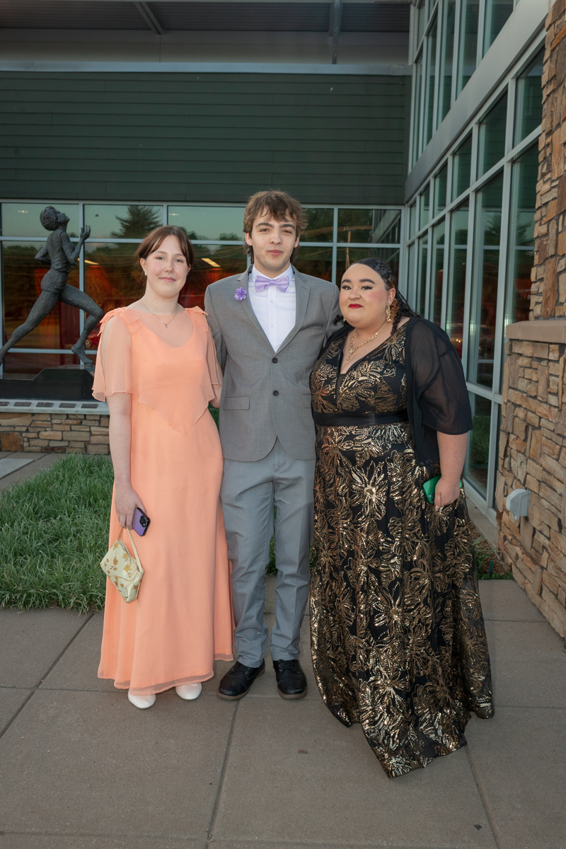 Three young adults in formal attire pose together outside a glass-and-stone building, with a peach dress on the left, a light gray suit and lavender bow tie in the center, and a black-and-gold sequined gown on the right.