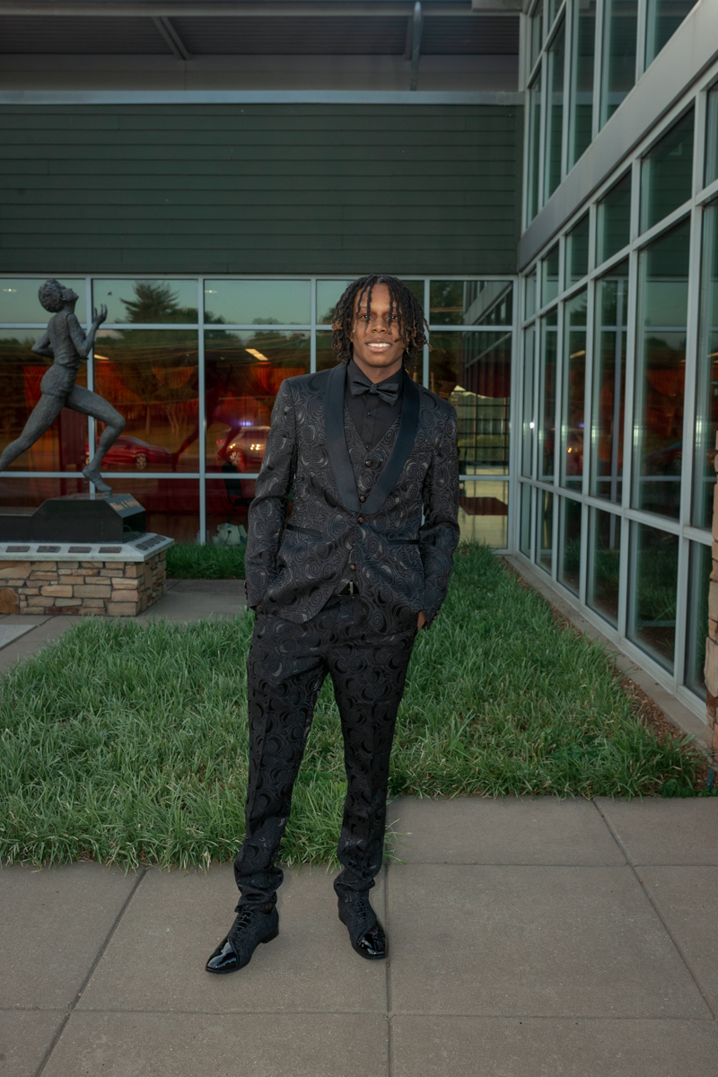 Young man in a black patterned suit with a bow tie standing on a sidewalk outside a glass-front building with a sculpture nearby.