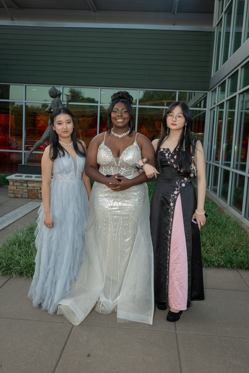 Three young women in formal gowns pose together outside a glass-front building; center wears a silver beaded gown with a flower corsage on her wrist, flanked by a pale blue gown on the left and a black/pink gown on the right.