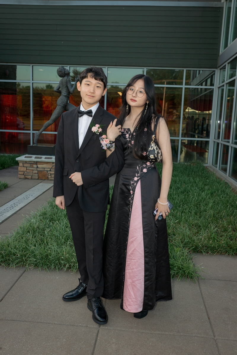 Couple in formal attire posing outdoors, man in a tuxedo and woman in a black-pink gown, arm in arm.