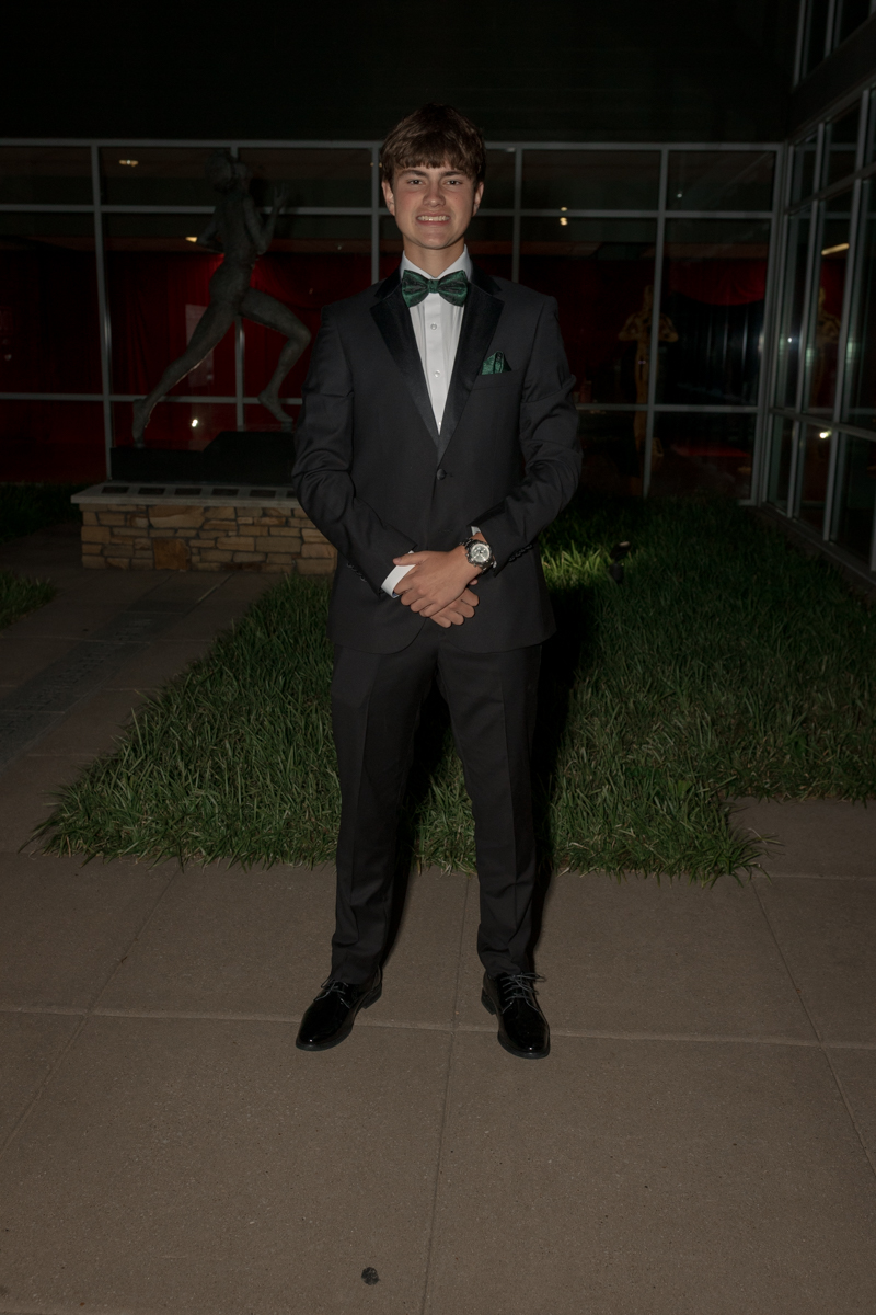 Young man in a black tuxedo with green bow tie and pocket square, posing outdoors at night by a statue and glass building behind him.