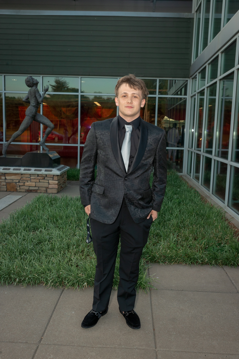 Young man in a dark patterned tuxedo with a silver tie standing outside a modern glass-front building, hands in pockets.