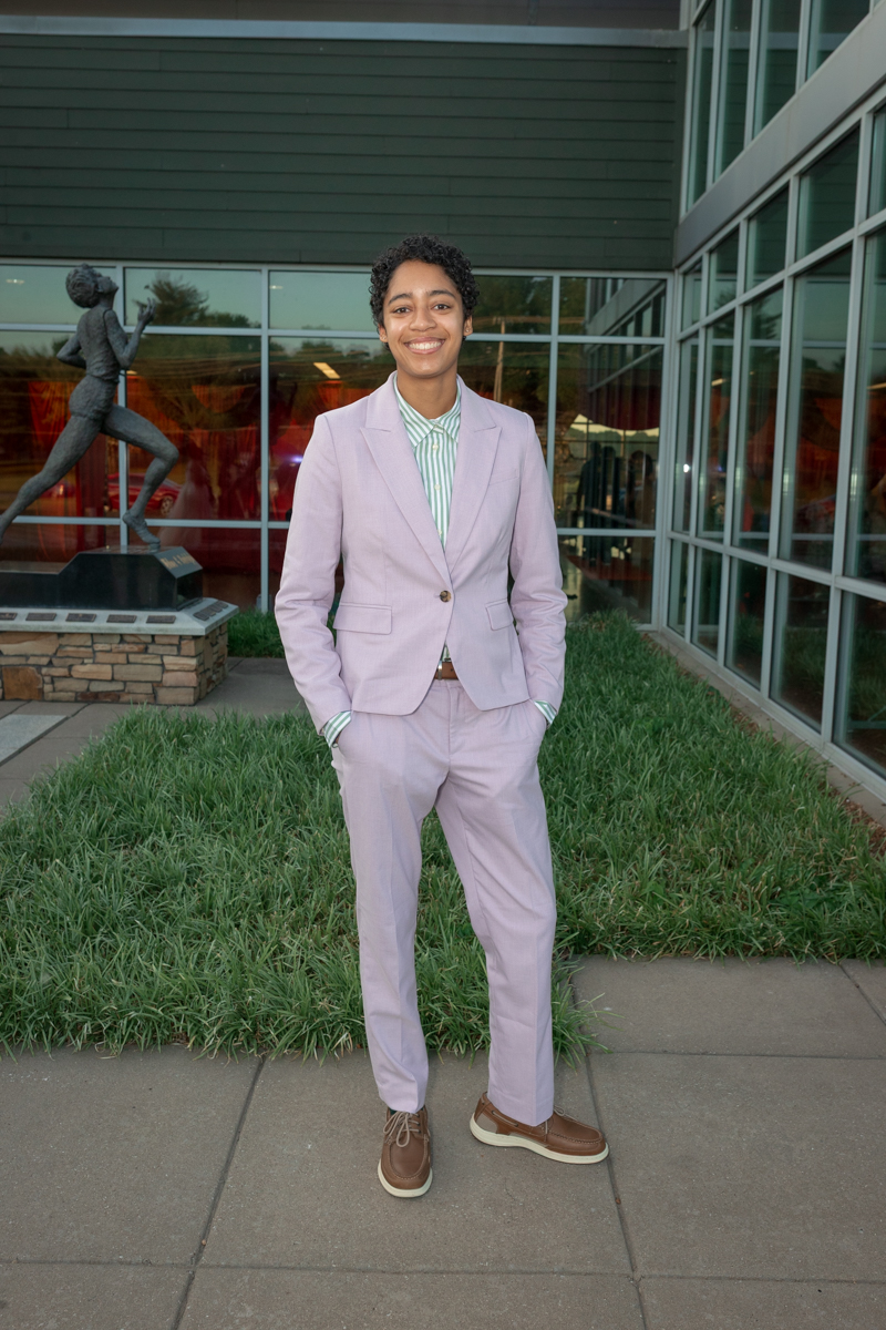 Person with short curly hair in a light purple suit standing outdoors near a modern glass building and a statue, smiling at the camera.