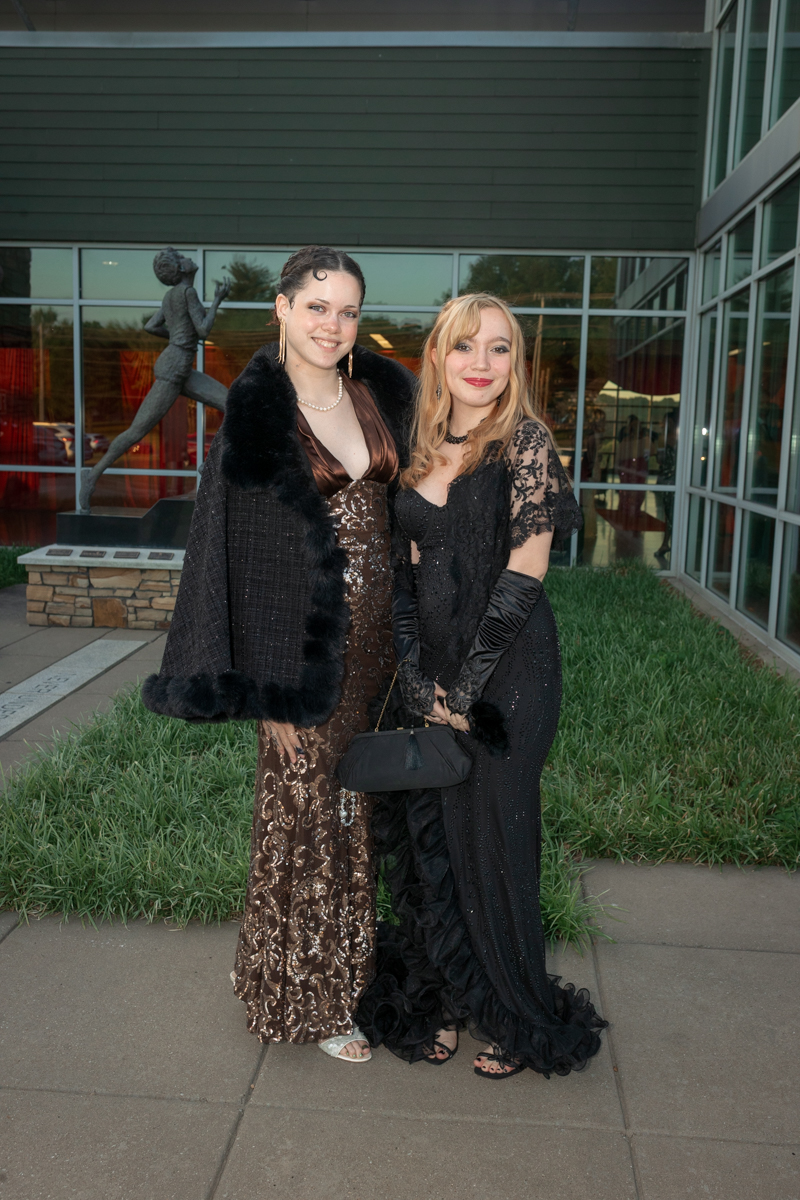 Two women in formal evening gowns pose together outside a glass-front venue, smiling for the camera, one in a brown sequined dress with a fur stole and the other in a black lace gown with gloves and a clutch.