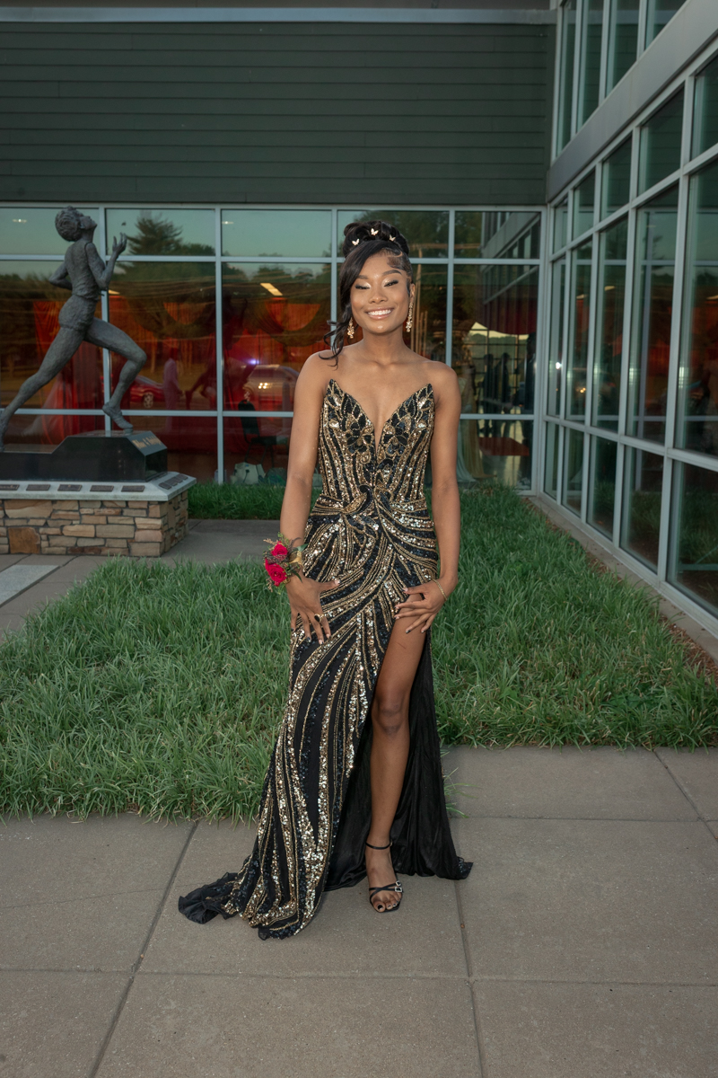 Young woman in a gold-and-black sequin evening gown with a thigh-high slit, posing outdoors in front of a modern glass building.