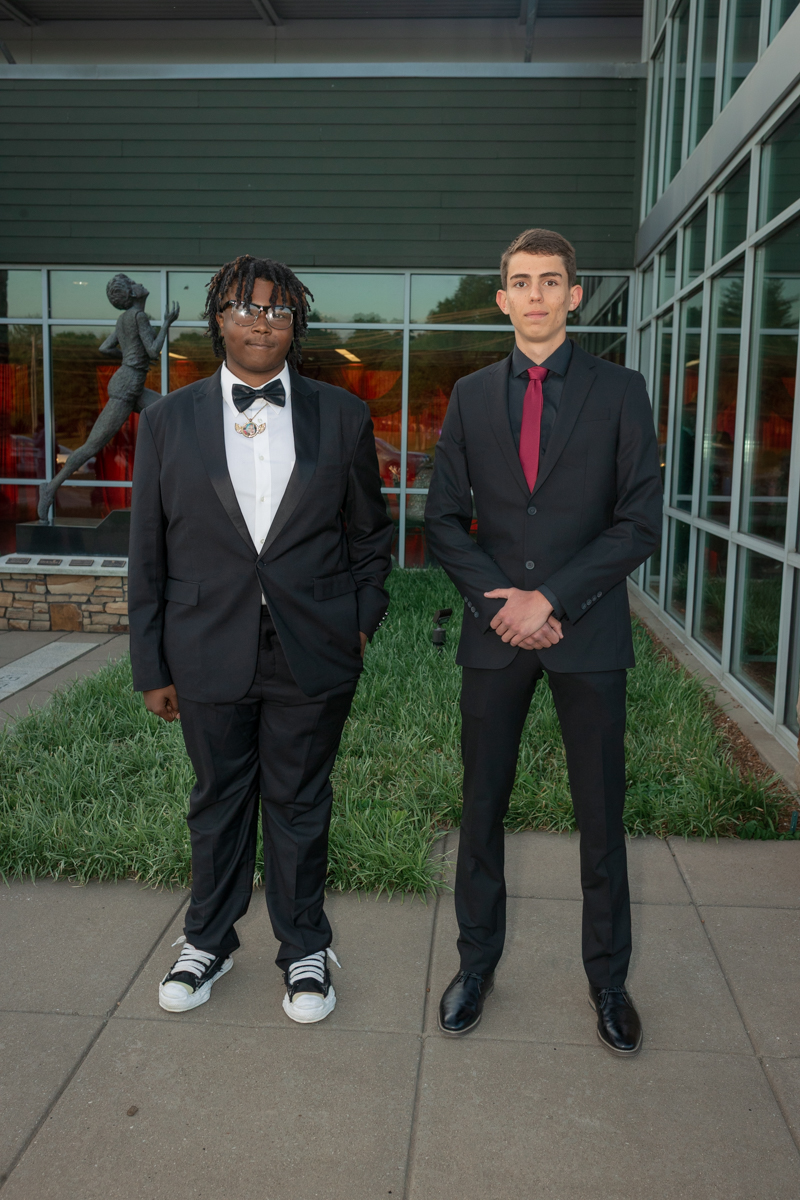 Two young men in formal suits pose outside a glass-front building; one in a tux with a bow tie and sneakers, the other in a dark suit with a red tie.