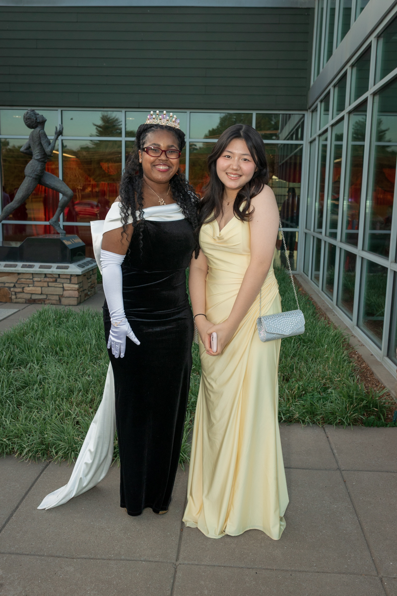 Two young women in formal gowns pose together outside a glass-front building; one wears a crown and a black velvet dress with white gloves, the other a pale yellow gown with a silver purse.
