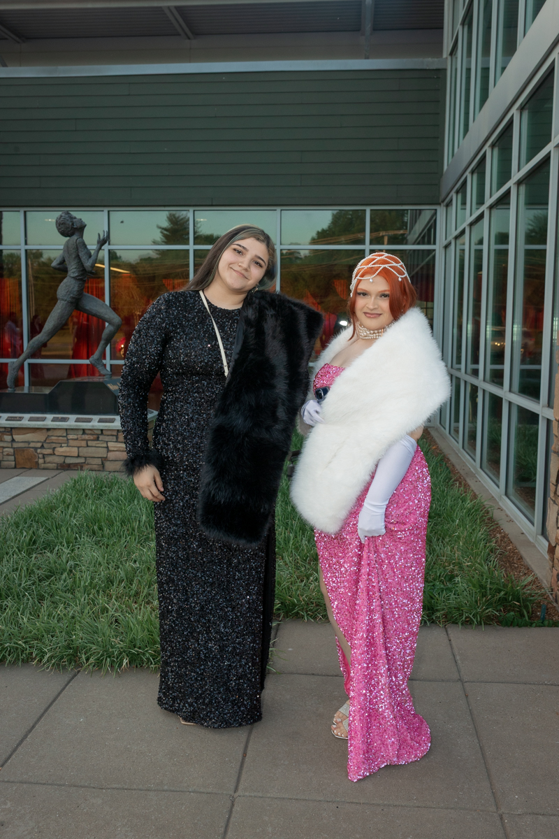 Two women in formal gowns posing outdoors: one in a black sequin dress with a black fur stole, the other in a pink sequin dress with a white fur wrap and decorative headpiece.