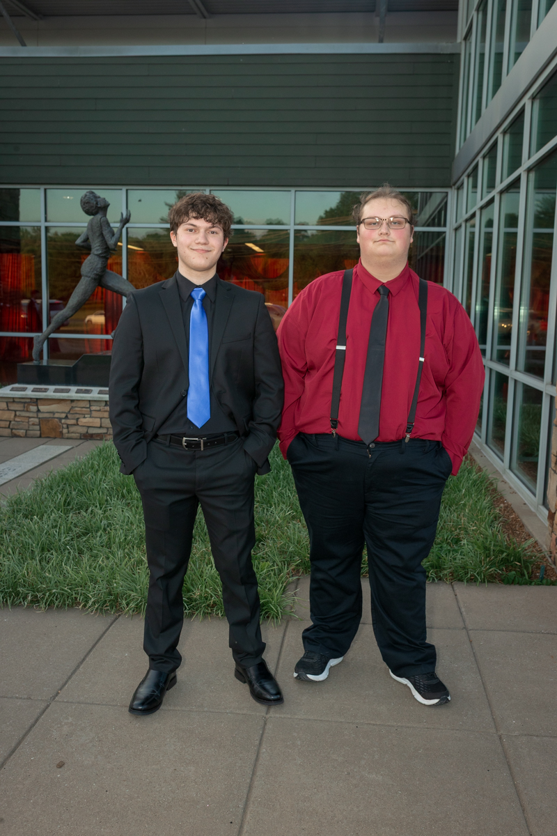 Two young men stand outside a modern building in formal attire: one in a black suit with a bright blue tie, the other in a red shirt with a black tie and suspenders.