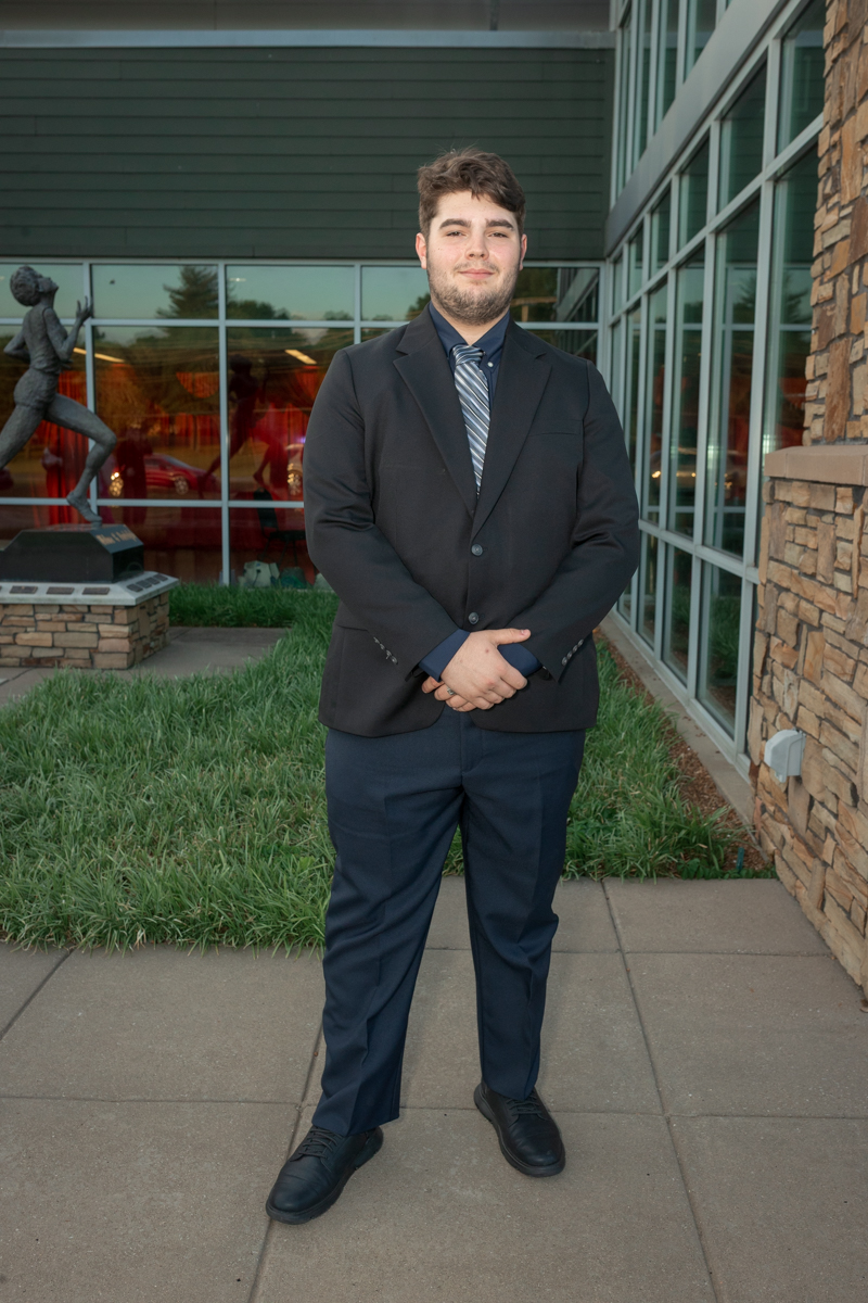 Young man in a dark suit and striped tie stands with hands clasped outside a modern glass-front building, near a statue and landscaping.