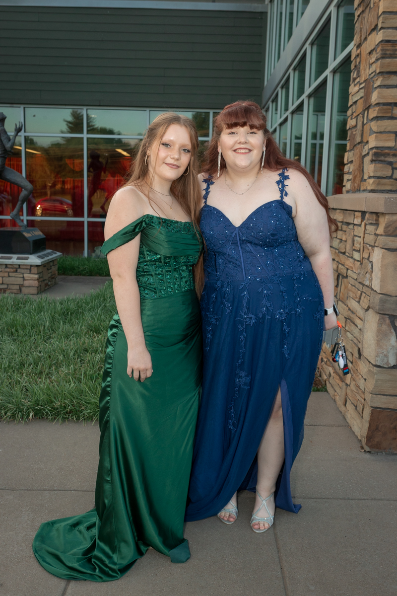 Two young women in formal gowns posing outside a building, one in a green off-shoulder dress and the other in a blue embellished gown with a slit, smiling at the camera.