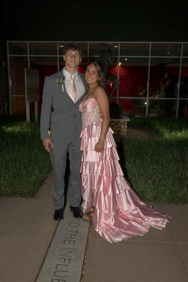 Prom couple posing outdoors at night: girl in a pink tiered satin gown and the boy in a gray suit with a pink tie, smiling at the camera.