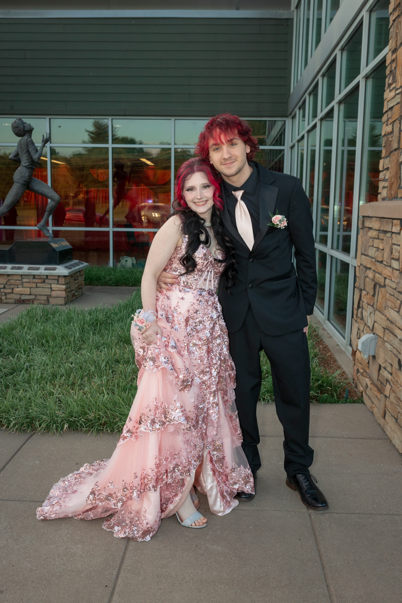 Two teenagers in formal attire pose outside a glass-fronted building, the girl wearing a pink sequined gown and the boy in a black suit with a pale pink tie.