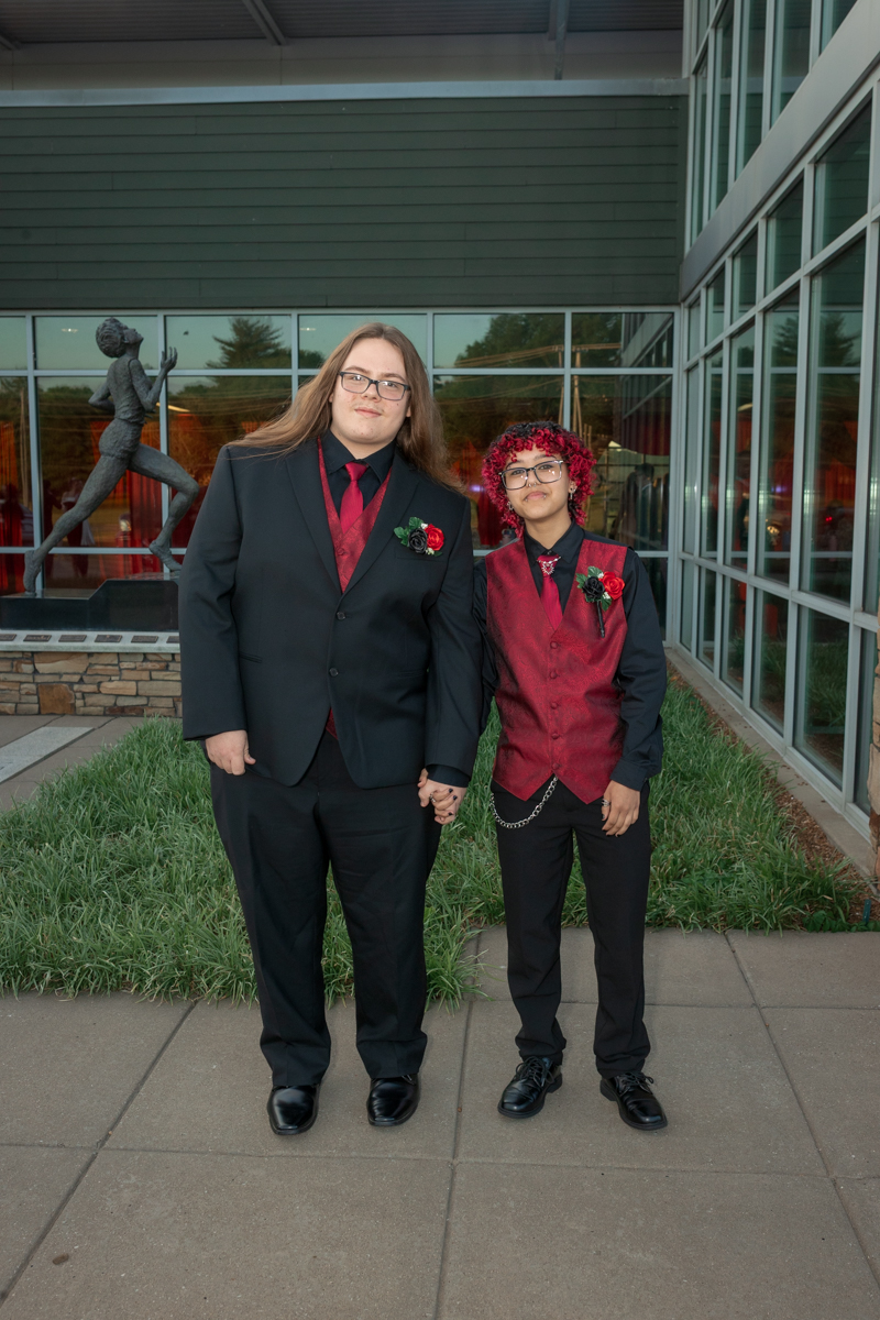 Two people in formal outfits stand outside a glass-front building, holding hands. They wear black suits with red vests and boutonnières, smiling at the camera.