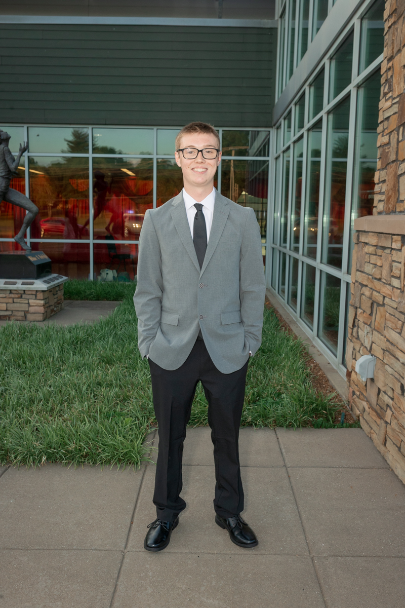 Young man in a gray suit, white shirt, and black tie stands outside a glass-front building with stone accents.