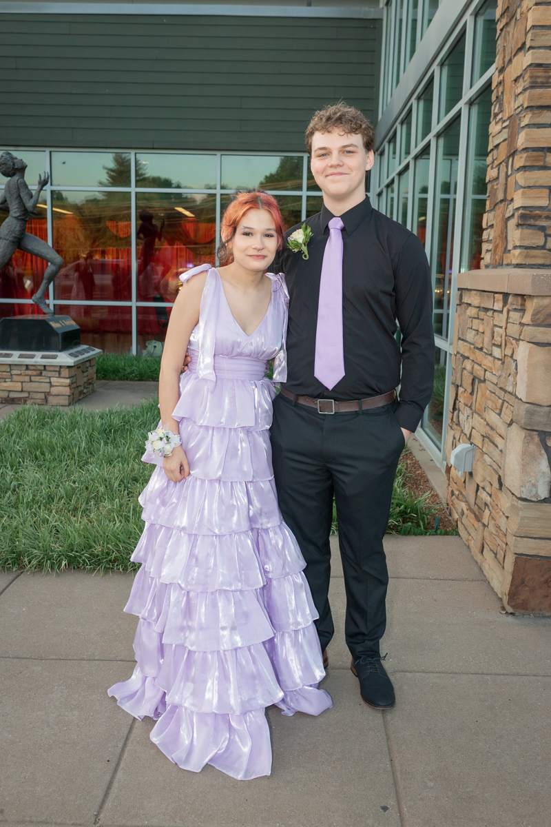 Girl in a lilac tiered gown and a boy in a black shirt with a lavender tie pose together outside a building.