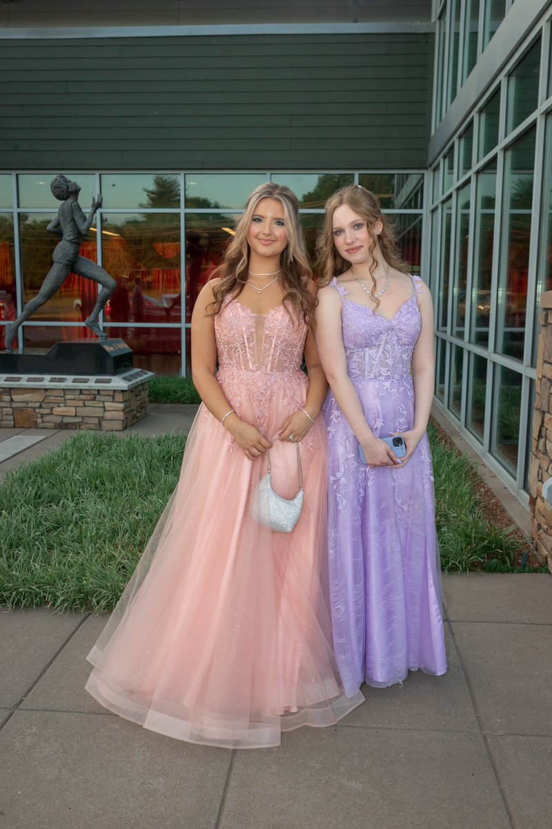 Two young women in formal gowns pose together outside a modern glass building, one in pink, the other in lavender, smiling at the camera.