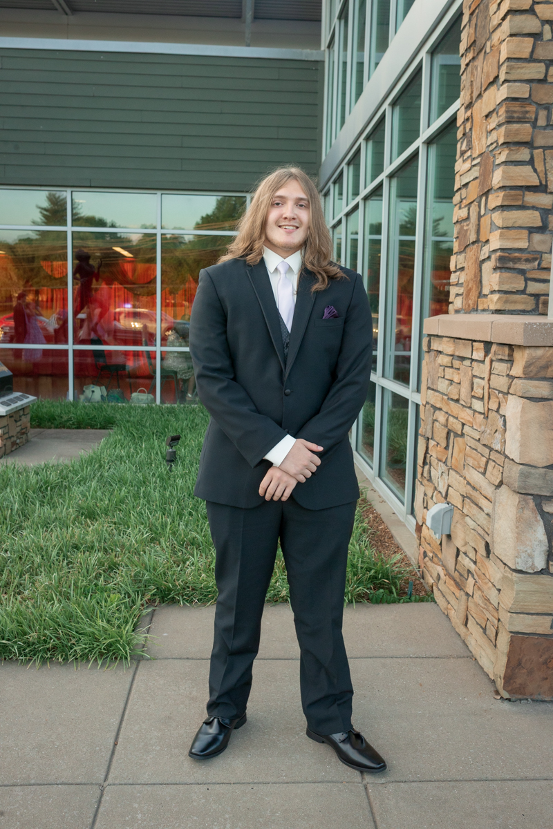 Man in a dark suit with a white shirt and tie standing outside a glass-fronted building with stone accents, hands clasped in front.