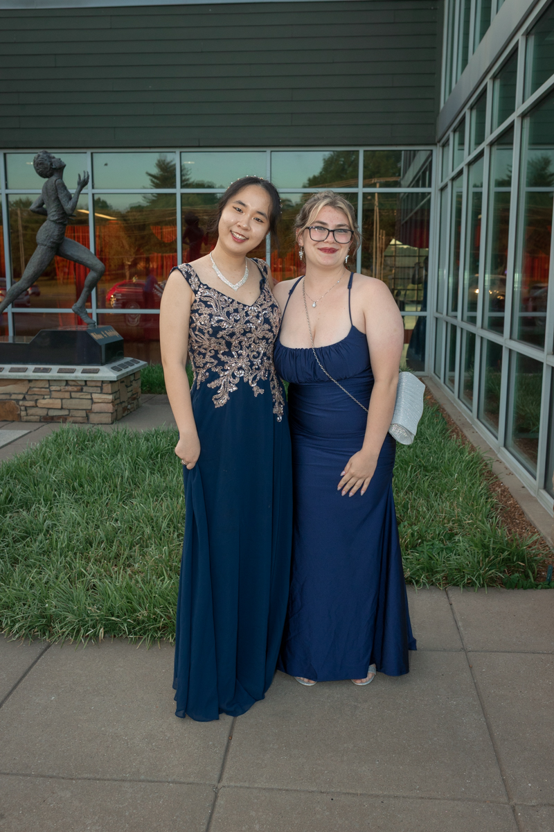 Two women in navy evening gowns pose together outside a glass-front building, one with gold embroidery on her dress.