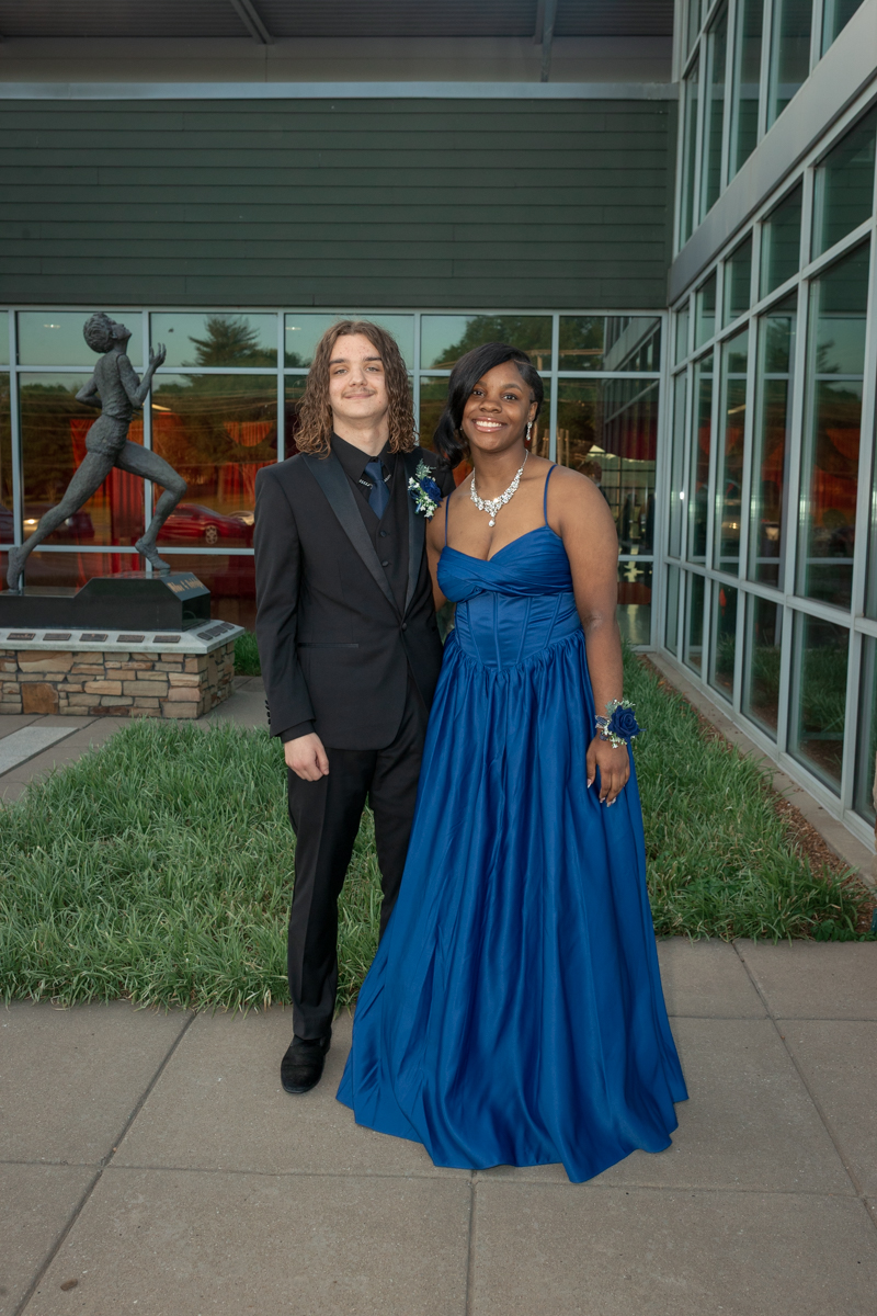 Two teens in formal wear pose together outside a glass-fronted building; boy in a black suit and tie, girl in a blue ball gown with a statement necklace and corsage on her wrist.