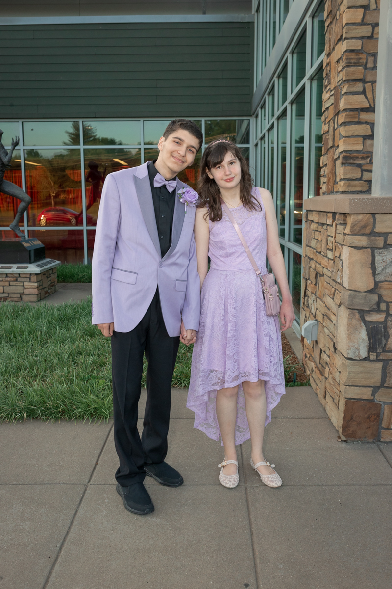 Two teens in lavender formal wear holding hands outside a glass-front building; boy wears a lavender suit with a bow tie, girl in a lavender lace dress with a pink crossbody bag.