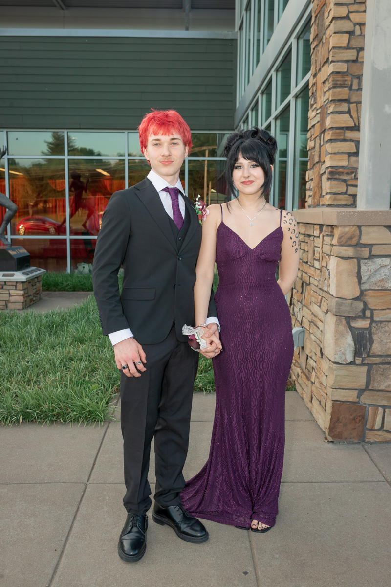 Couple dressed for a formal event posing outside a modern building, holding hands; man in a black suit with a purple tie and red hair, woman in a purple evening gown.