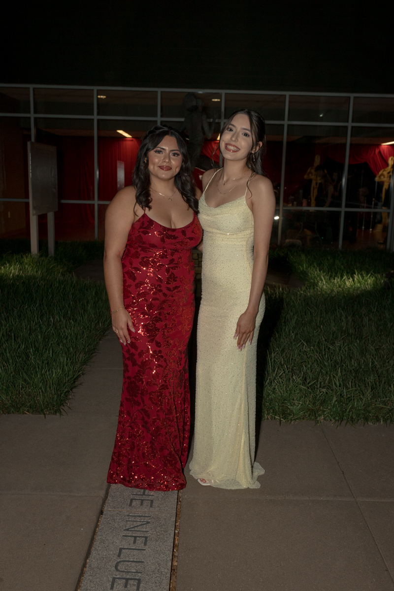 Two young women in formal gowns pose together outdoors at night, smiling for the camera, one in a red sequined dress and the other in a pale gold dress.