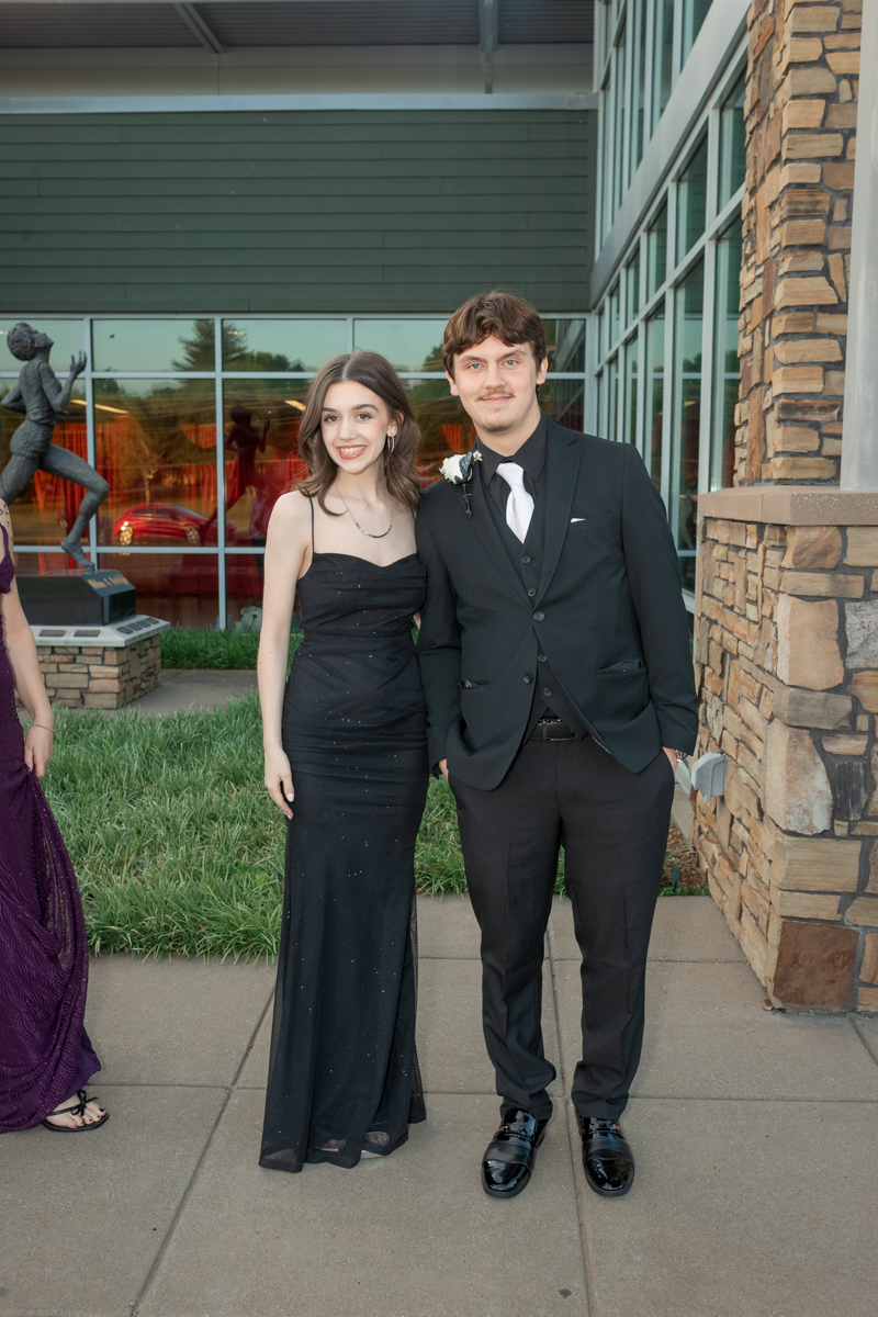 Two people in formal attire posing outside a modern brick-and-glass building; a girl in a black strapless gown and a boy in a black suit with a white tie and boutonniere stands side by side.