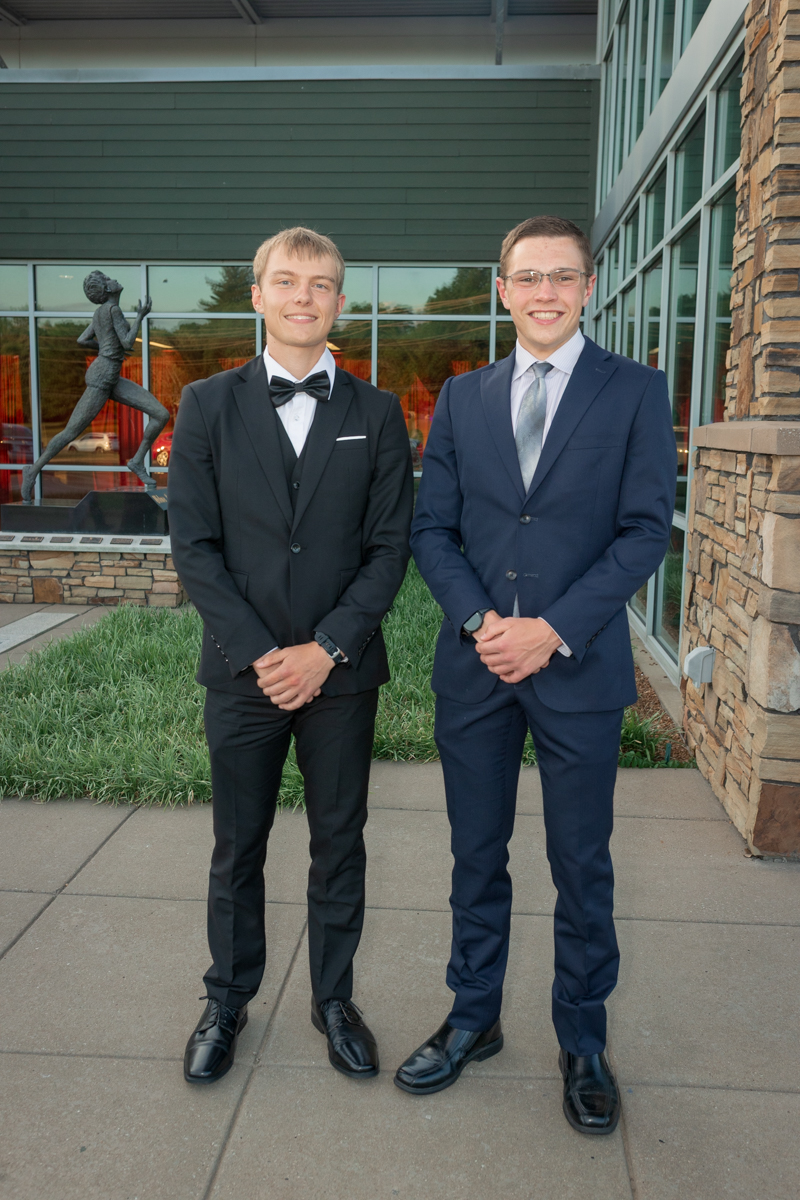 Two young men in formal attire stand side by side outside a building, smiling; one in a black tux with bow tie, the other in a navy suit with light gray tie.